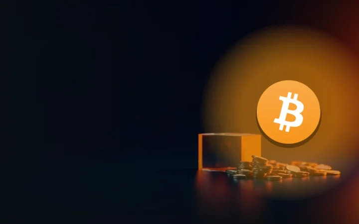 Bitcoin-logoet over en mørk navyblå baggrund med glødende orange coins og en glasblok til højre.