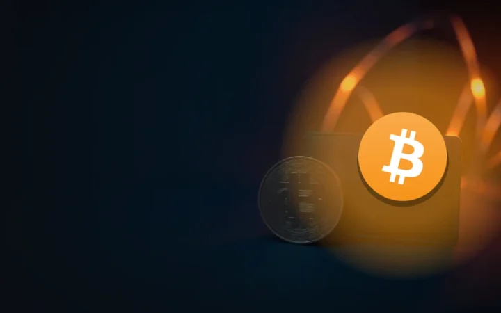 Bitcoin-logoet over en blank metalmønt og en skærmløs sort wallet på et mørkt navyblåt skrivebord med orange glødende lysspor i højre side.