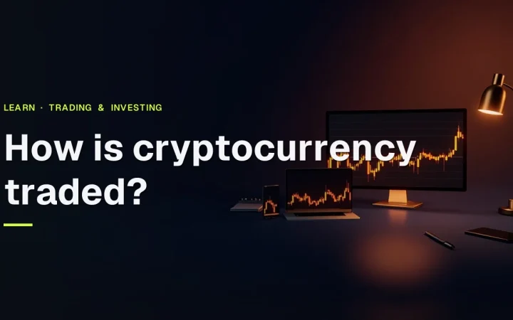 The title How is cryptocurrency traded? over a dark desk with a laptop, phone and monitor showing glowing orange charts.