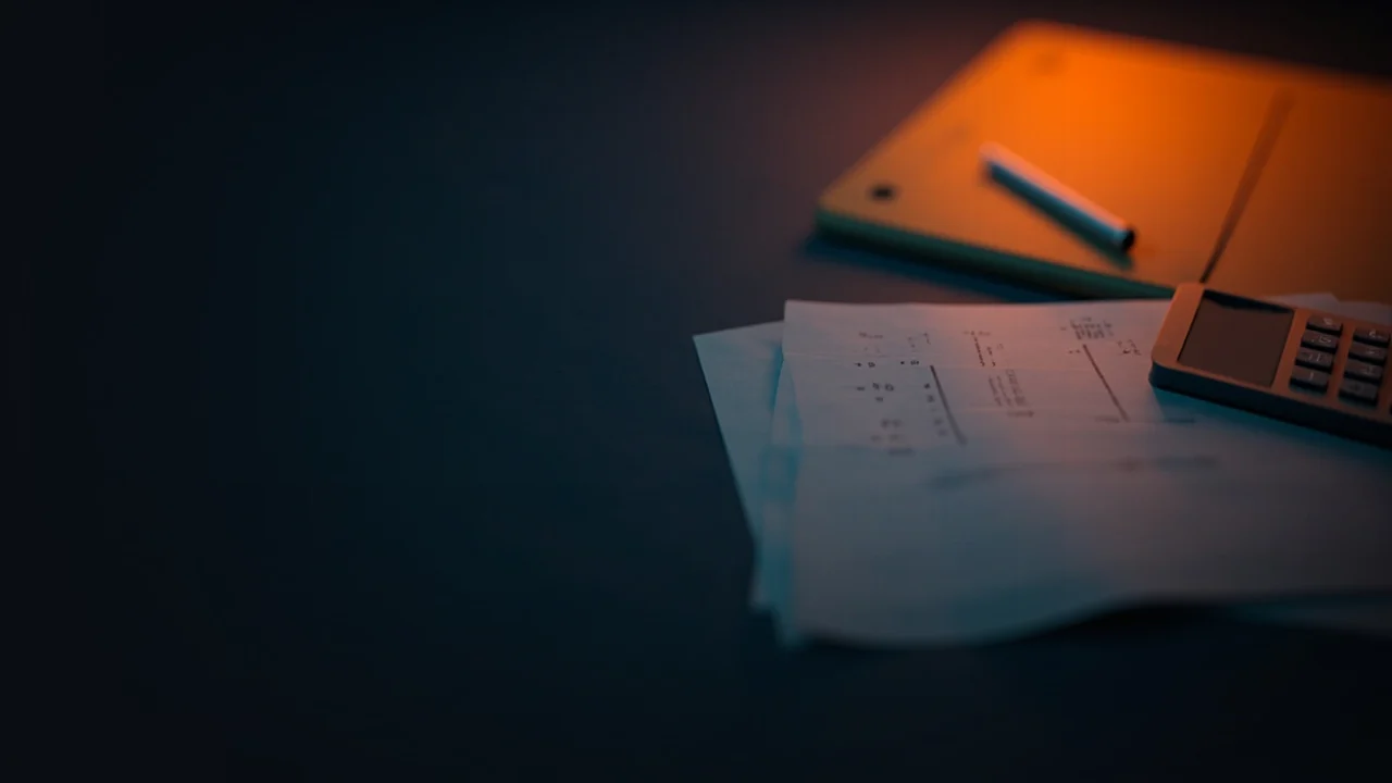 Ein dunkler Schreibtisch mit leeren Steuerformularen, einem Taschenrechner, einem Stift und einem Ordner in warmem orangefarbenem Licht.