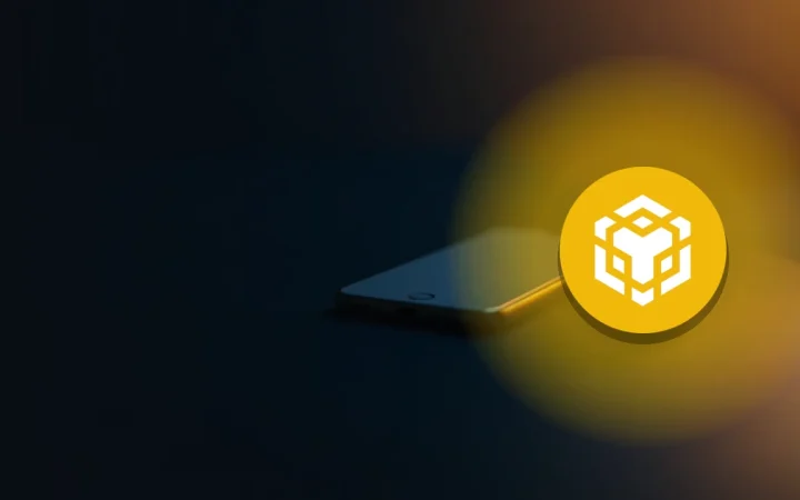 The BNB logo over a dark desk holding a smartphone and coins.