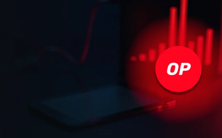 El logotipo de Optimism sobre un escritorio oscuro con un dispositivo de cartera, un cable rojo brillante y un monitor que muestra barras rojas.