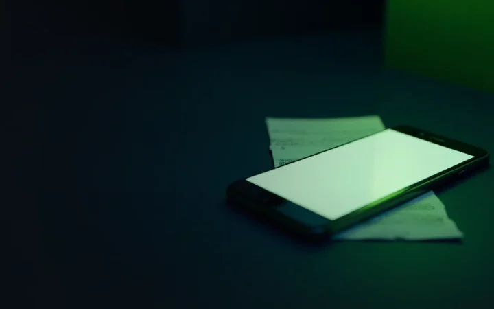A dark desk with a blank smartphone and a folded bank statement on the right, lit by lime green light, with the left side empty and dark.