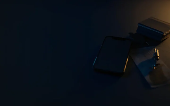 Un bureau bleu marine sombre avec un smartphone vierge, des cartes, une clé et du papier éclairés par des accents dorés, côté gauche vide et ombragé.