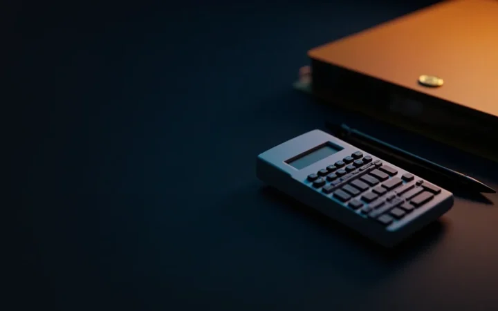 A blank calculator, pen and folder on a dark desk under orange light.