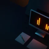 A dark desk scene with a blank monitor, notepad and pen, lit in orange.