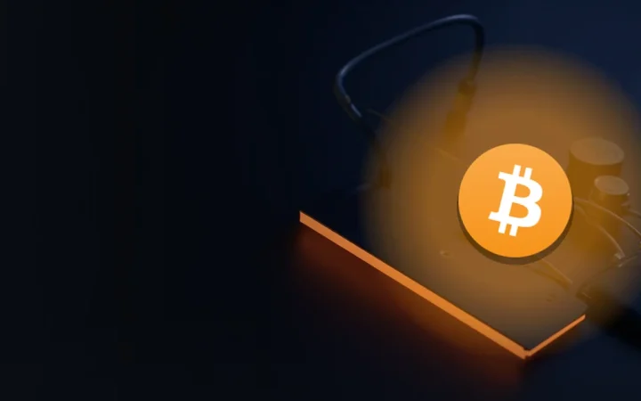 Bitcoin-logoet over et mørkt skrivebord med et glødende kabel og en enhed med tom skærm.
