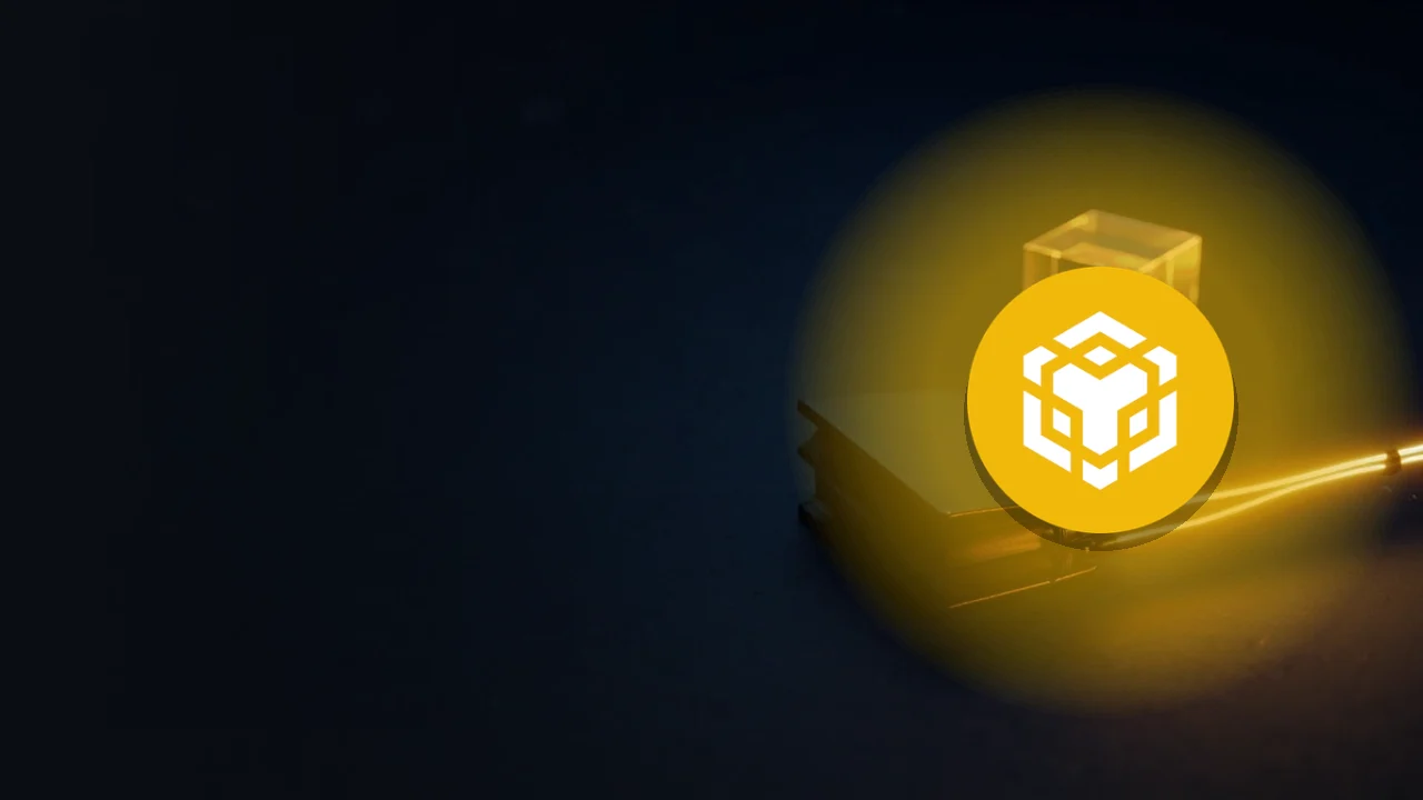 Das BNB-Logo über einem dunklen Schreibtisch mit einer Wallet und leuchtenden Goldkabeln.