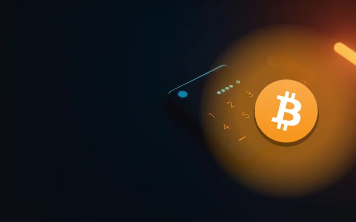 暗いネイビーのデスク面に置かれた、すっきりとした電卓のようなデバイスの上に重なるBitcoinのロゴ。空白のディスプレイが柔らかく光っている。