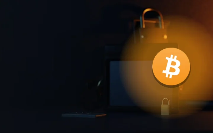 The Bitcoin logo over a dark desk with a hardware wallet, steel plate, padlock, and shield lit in orange.