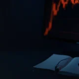 A dark desk with a glowing candlestick chart and a closed notebook.